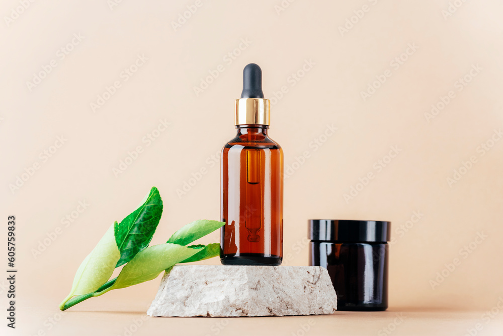 Brown cosmetic serum bottle on natural stone podium and cream jar, green leaves on neutral beige background. Closeup Brown cosmetic serum bottle on natural stone podium and cream jar, green leaves on neutral beige background. Closeup