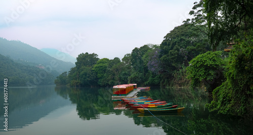 In this picturesque landscape photo, Phewa Lake in Pokhara, Nepal, unfolds its serene beauty. The calm waters of the lake reflect the surrounding mountains, creating a breathtaking panorama. A cluster (ID: 605774413)