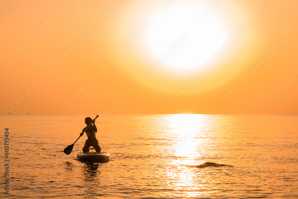 Girl wearing sun glasses and white bikini poses sitting on a sup board and oar using paddle