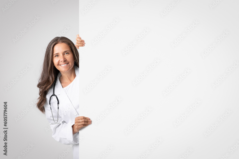 Smiling senior woman medical worker looking out behing white advertising board with free space for text or design Smiling senior woman medical worker looking out behing white advertising board with free space for text or design