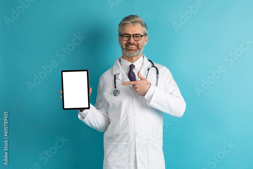 Cheerful mature doctor showing digital tablet with mockup on blue