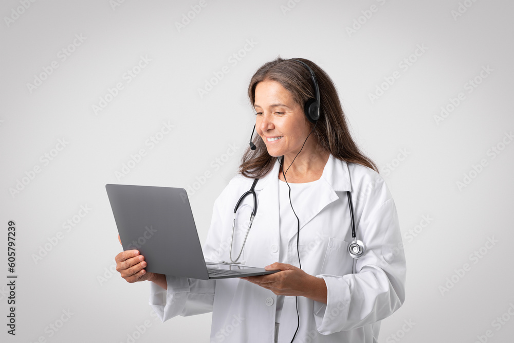 Cheerful european senior woman doctor in white coat with stethoscope, headphones video calling on laptop Cheerful european senior woman doctor in white coat with stethoscope, headphones video calling on laptop