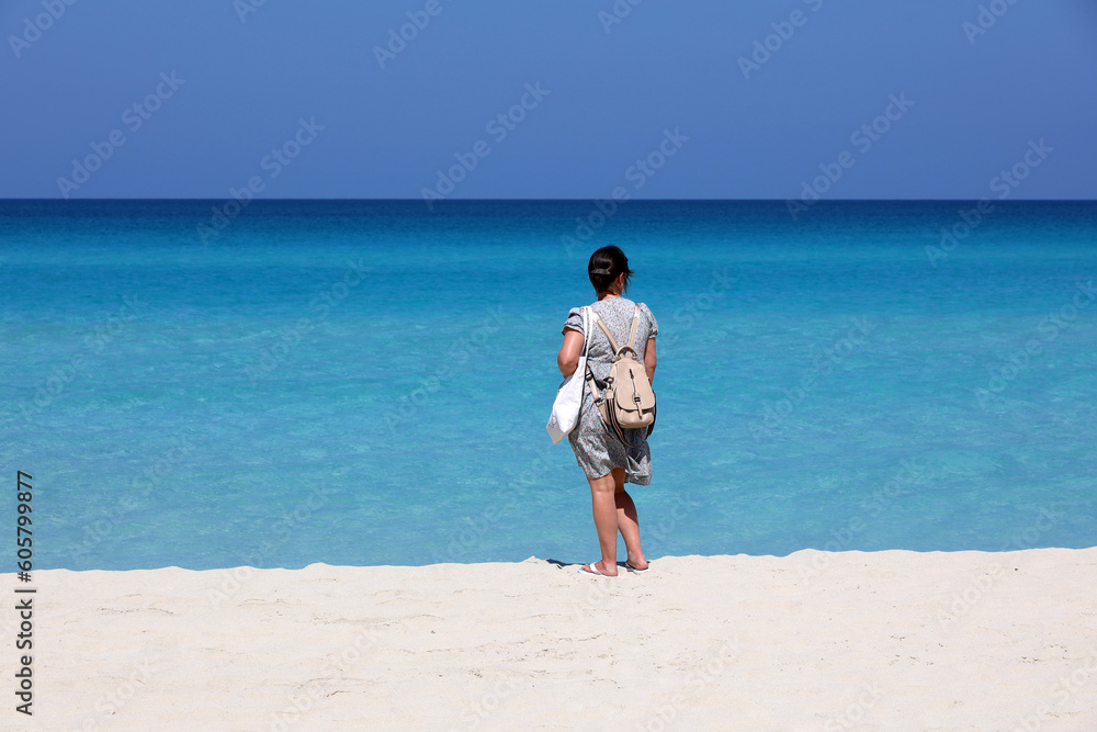 Woman in summer dress standing with backpack purse on white sand against the azure waves. Travel and beach vacation on ocean coast