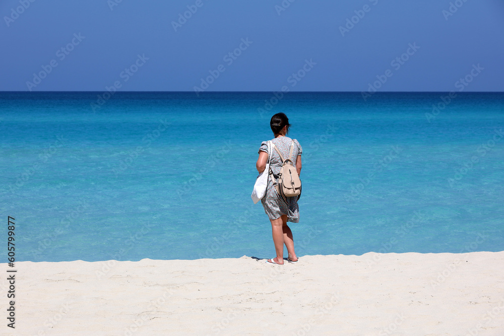 Woman in summer dress standing with backpack purse on white sand against the azure waves. Travel and beach vacation on ocean coast Woman in summer dress standing with backpack purse on white sand against the azure waves. Travel and beach vacation on ocean coast