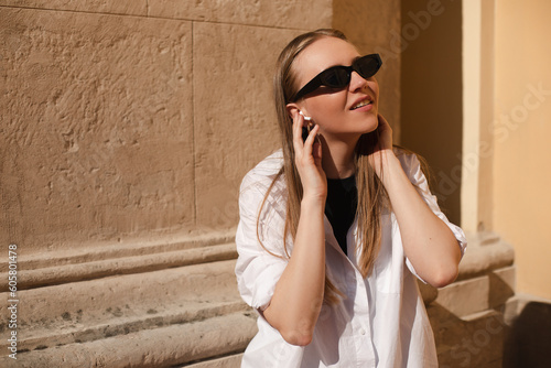  Pensive lady in stylish white shirt and black tot  glasses enjoying of good music. Outdoor portrait of blonde girl in white wireless earphones listening music walking near wall. Touch headphones.