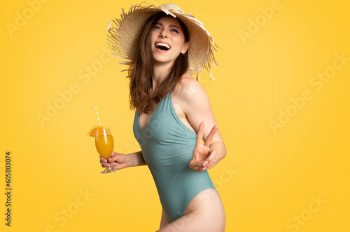 Relax at vacation on beach. Excited woman in swimsuit and straw hat enjoying cocktail, showing peace sign and smiling #605803074