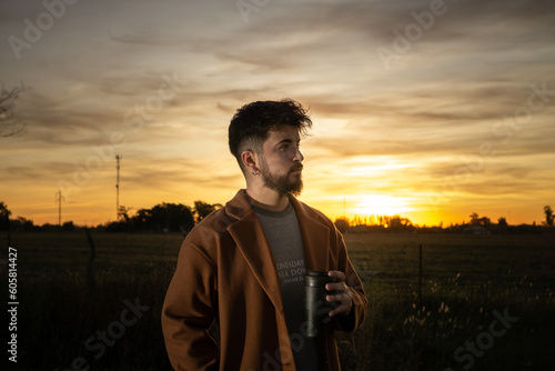 young man drinking coffee in the field with a sunset in the background © Eric