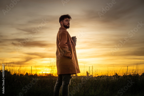 young man drinking coffee in the field with a sunset in the background