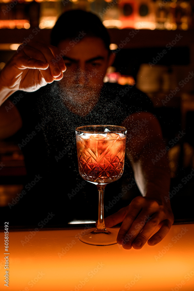 Barman sprinkle glass with alcoholic cocktail with piece of orange citrus zest