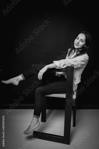 emotional portrait of cute and seductive brunette girl sitting in pants and white shirt on chair isolated on dark background