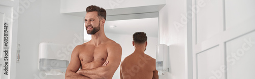 Positive man with towel and crossed arms leans onto cabinet near mirrror in bathroom photo