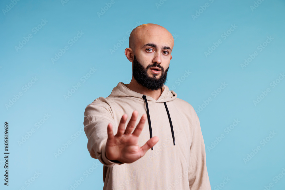 Confident arab man making stop gesture with hand, expressing rejection portrait. Young person in casual hoodie showing no signal with palm and looking at camera with serious emotions Confident arab man making stop gesture with hand, expressing rejection portrait. Young person in casual hoodie showing no signal with palm and looking at camera with serious emotions