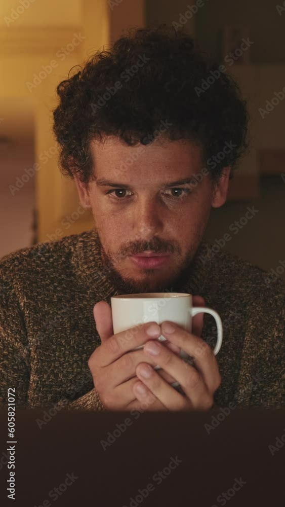 Young man sits in an apartment, drinks coffee from mug, uses laptop Young man sits in an apartment, drinks coffee from mug, uses laptop