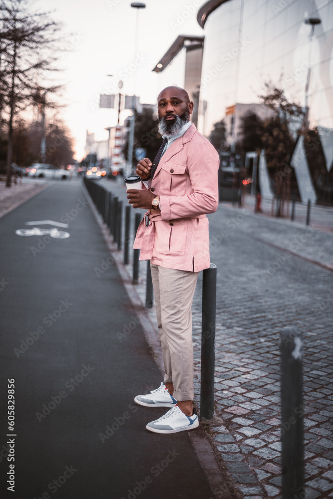 A stylish black bald man, with a long beard with grey hair, casually dressed in pink, holds a cup of coffee as he is about to cross the bicycle lane in the afternoon, he checks if the road is clear A stylish black bald man, with a long beard with grey hair, casually dressed in pink, holds a cup of coffee as he is about to cross the bicycle lane in the afternoon, he checks if the road is clear