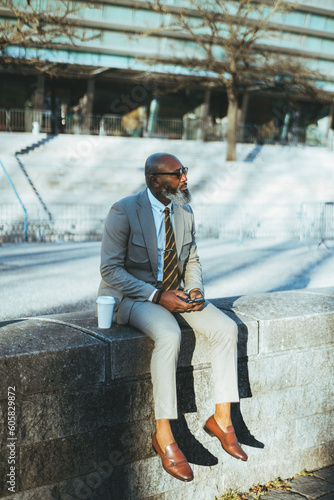 A bald man with a full white-bearded face holding a smartphone, has his cup of coffee next to him, dressed in a cream suit with a yellow striped tie, sitting on limestone steps wearing sunglasses