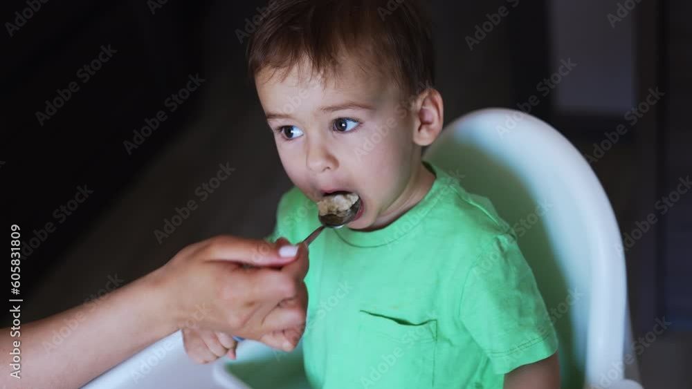 Toddler sitting at feeding table eats from fork. Mom’s hand gives a spoon with porridge to her child. Top view close up. Toddler sitting at feeding table eats from fork. Mom’s hand gives a spoon with porridge to her child. Top view close up.