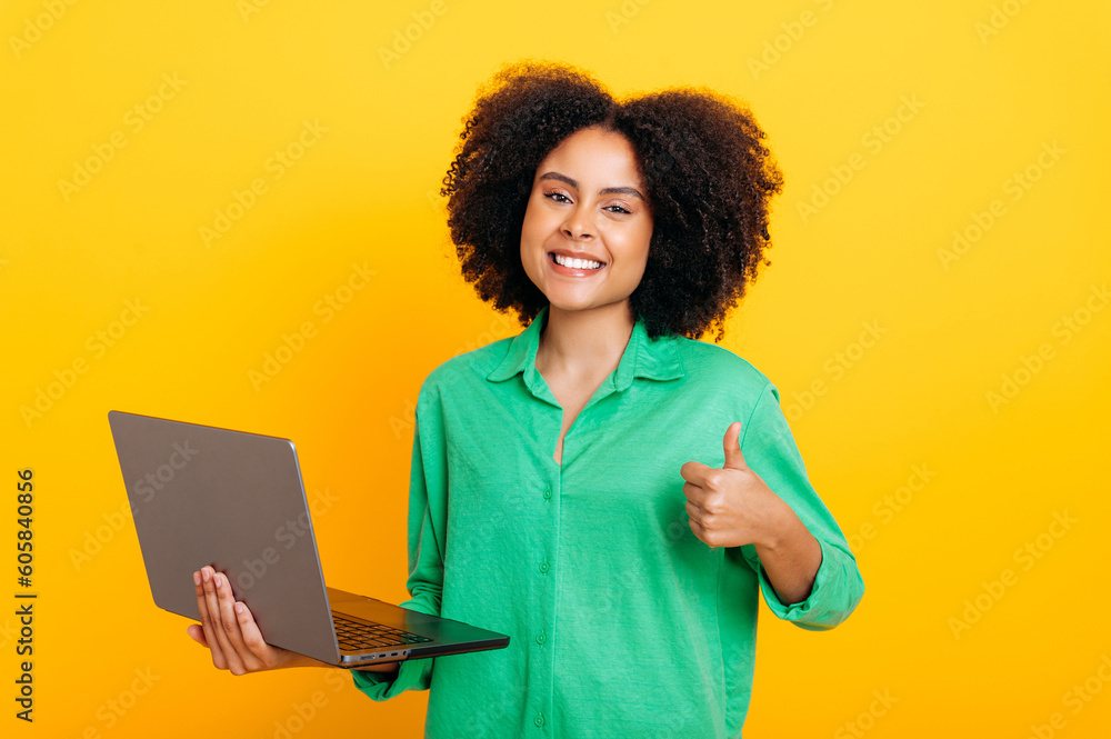 Joyful gorgeous brazilian or african american curly haired woman, in a green shirt, holding an open laptop in hand, looks at camera, smiles, shows thumb up gesture, stand on isolated orange background Joyful gorgeous brazilian or african american curly haired woman, in a green shirt, holding an open laptop in hand, looks at camera, smiles, shows thumb up gesture, stand on isolated orange background