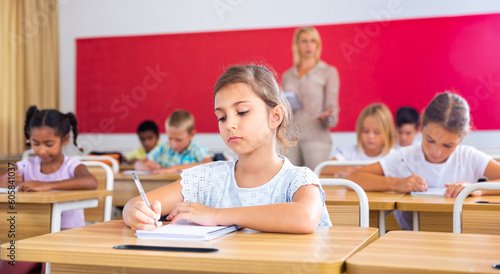 Attractive smart preteen girl studying in classroom, listening to teacher and writing in notebook