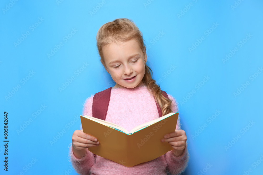 Happy little girl with backpack reading book on light blue background Happy little girl with backpack reading book on light blue background