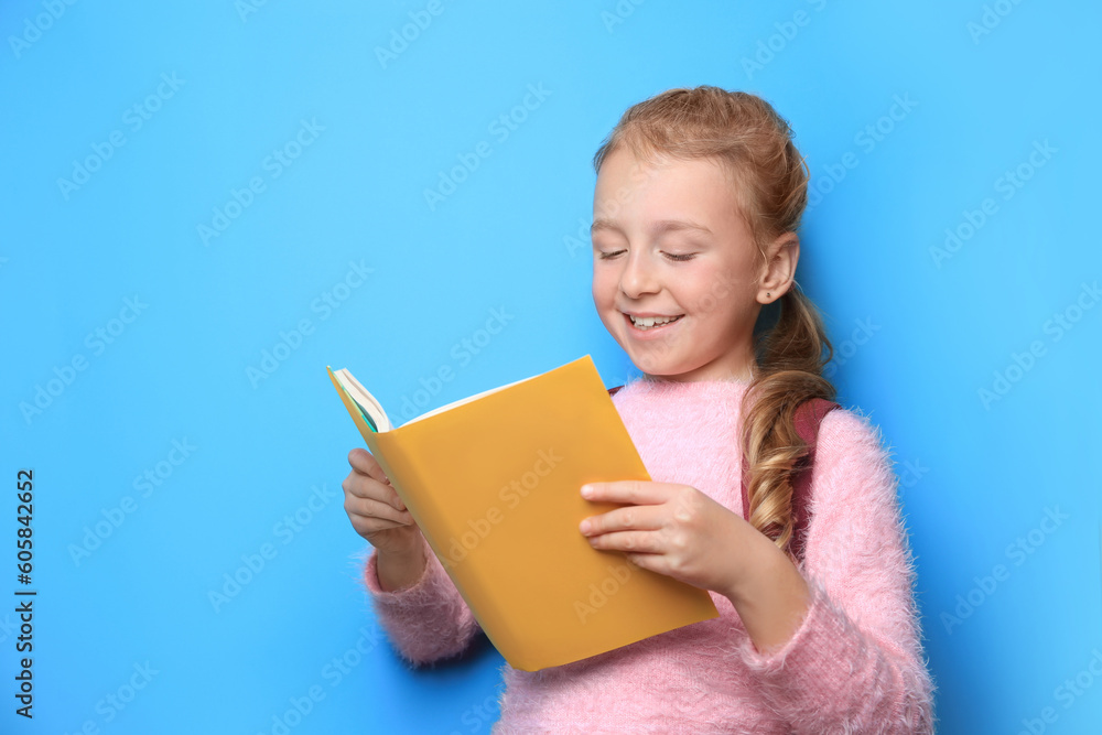 Happy little girl with backpack reading book on light blue background Happy little girl with backpack reading book on light blue background