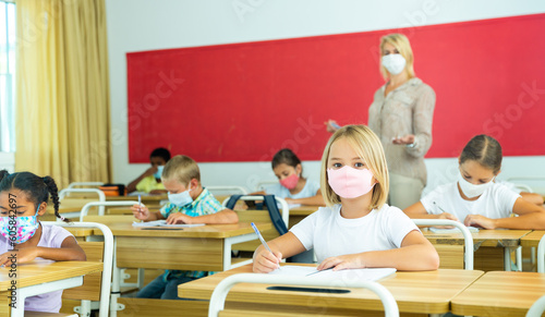 Primary school students in protective masks sit at school desk in the classroom