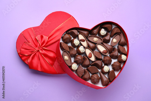 Heart shaped box with delicious chocolate candies on lilac background, top view