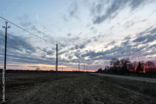Country road and local power line in a spring field with last year's grass on the outskirts of the settlement, blue hour at sunset. Beautiful sky with clouds in pink colors on the horizon, HDR