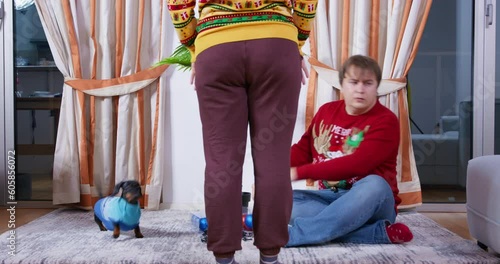 Man with dog in ugly sweater are sitting on floor near palm tree looking at festive balls. Grumpy wife drives them away with gesture of displeasure, throws decorations. Family preparing for Christmas photo