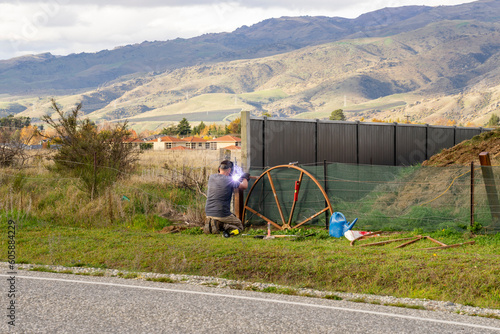 Man diligently welds metal construction in an outdoor setting. Surrounded by fields, mountains, and a sky adorned with clouds,
