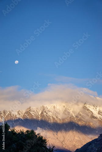 Vertical shot of snowy mountain range with a full moon in the clear dip blue sky.
