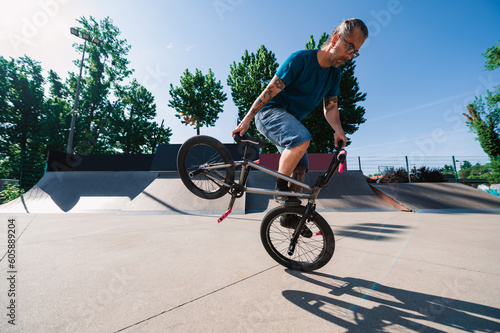 An oldish tattooed professional bmx bicyclist is doing freestyle tricks and stunts in a skate park.