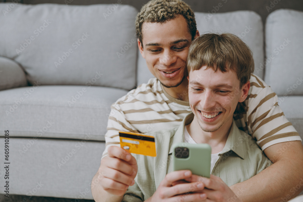 Young couple two gay men wear casual clothes use mobile cell phone credit bank shopping online sit on sofa couch home flat rest spend free time in living room. Pride day june month love lgbtq concept. Young couple two gay men wear casual clothes use mobile cell phone credit bank shopping online sit on sofa couch home flat rest spend free time in living room. Pride day june month love lgbtq concept.