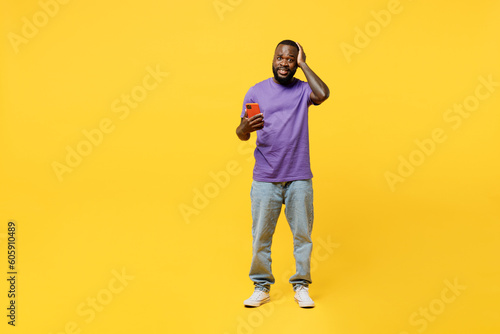 Full body young man of African American ethnicity wear casual clothes purple t-shirt hold head use mobile cell phone lok camera isolated on plain yellow background studio portrait. Lifestyle concept.
