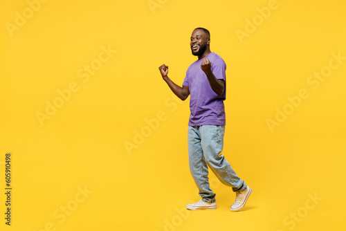 Full body sideways young man of African American ethnicity wear casual clothes purple t-shirt walk go doing winner gesture celebrate clenching fists say yes isolated on plain yellow background studio.