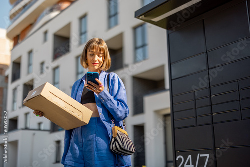 Young woman using smart phone while standing with a parcel delivered with post office machine with automatic lockers. New technologies in delivery service, self picking