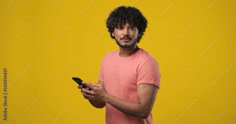 Young Smiling pensive indian man using mobile cell phone typing sms message looking aside up on orange studio background Young Smiling pensive indian man using mobile cell phone typing sms message looking aside up on orange studio background