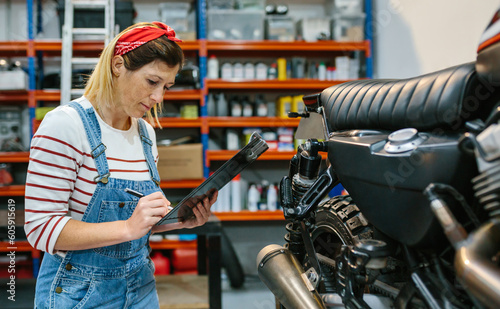 Concentrated mechanic woman with clipboard checking motorcycle and taking notes on factory