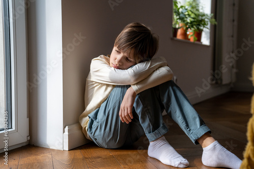Upset boy adopted child sitting alone on floor at home, feeling sad and lonely, suffering from anxiety and depression. Schoolboy kid feeling unhappy and depressed during puberty