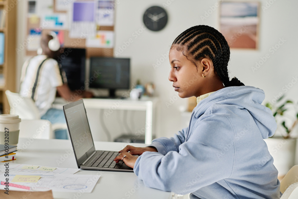 Side view of programmer with elegant hairstyle working on laptop sitting at her workplace in office