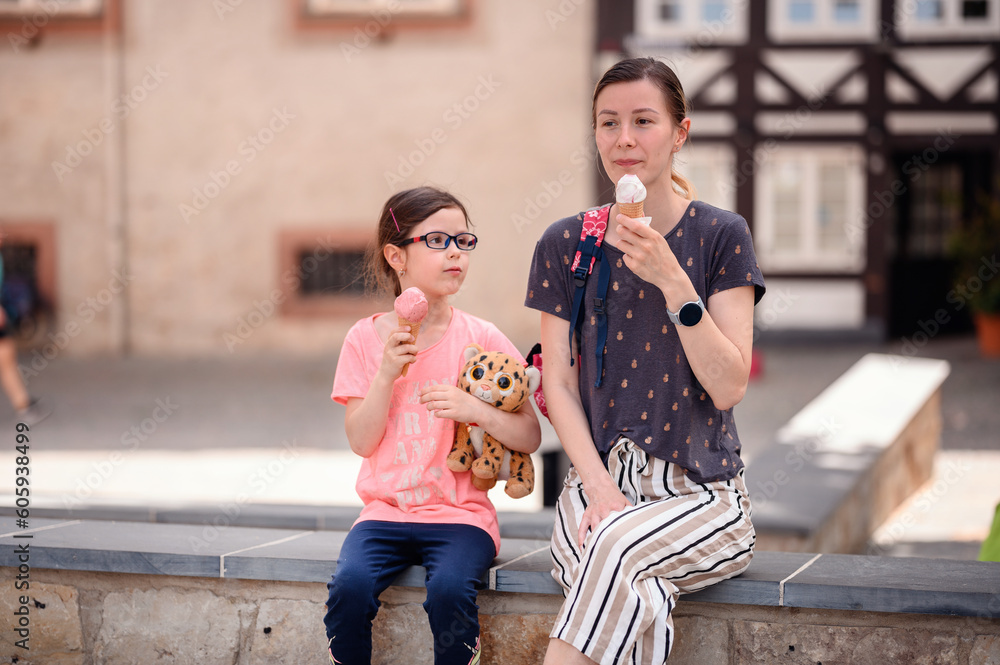 Young woman in summer outfit sits with little preschooler girl in glasses on stone wall
