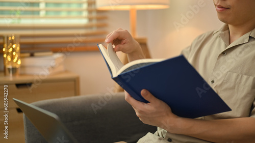 Calm young asian man reading interesting book on sofa in cozy home interior