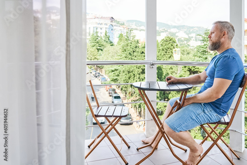 Bearded man sitting at wooden table on hotel balcony enjoys summer vacation. Male tourist explores sunny tropical resort town from room balcony