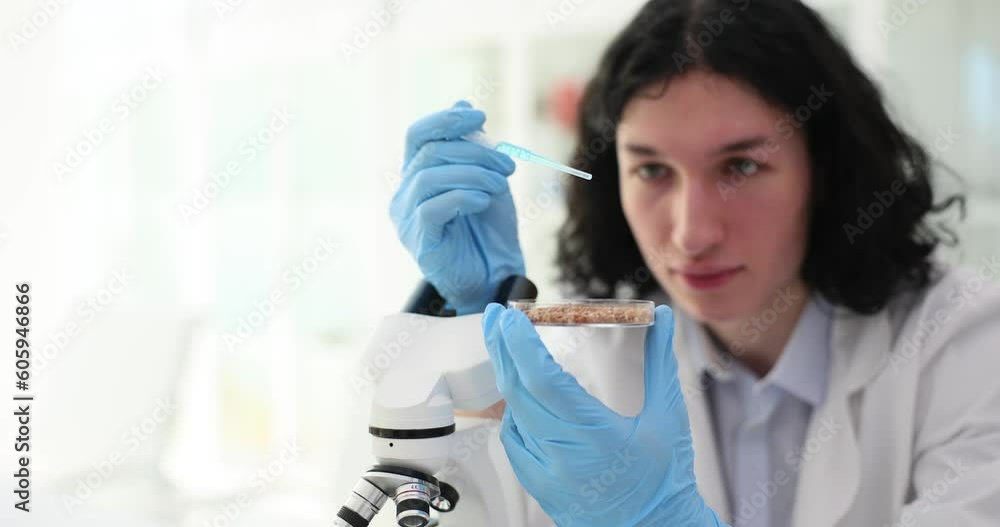 Man in rubber gloves holds pipette dripping light blue liquid reagent into Petri dish with seeds. Scientist uses equipment in agricultural laboratory Man in rubber gloves holds pipette dripping light blue liquid reagent into Petri dish with seeds. Scientist uses equipment in agricultural laboratory