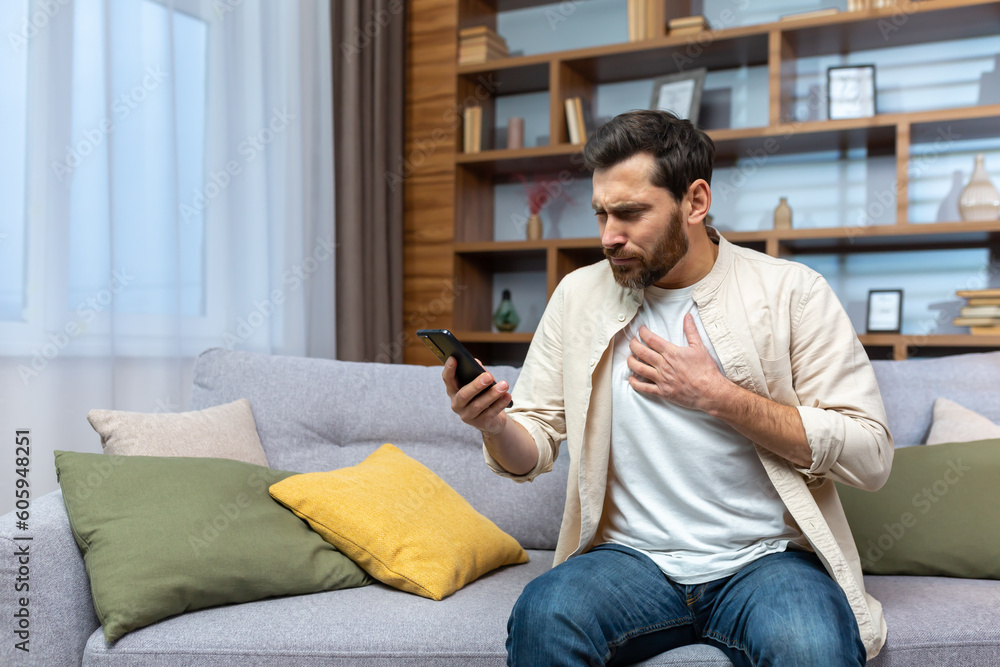 Mature man with beard alone at home reading bad news online on phone while sitting on sofa in living room at home.