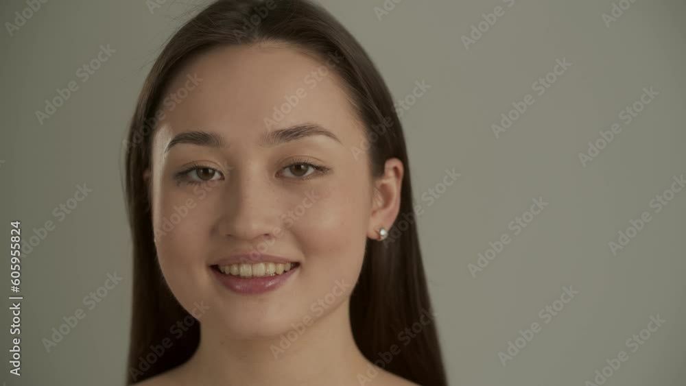 Pretty Asian woman posing against a gray wall. A young long-haired brunette stands on a gray background and smiles while looking at the camera. Pretty Asian woman posing against a gray wall. A young long-haired brunette stands on a gray background and smiles while looking at the camera.