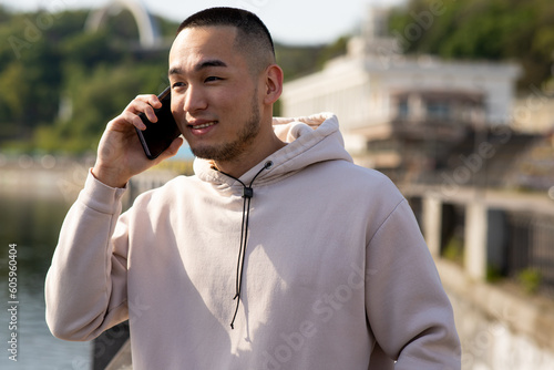 Happy Asian guy in sport hoodie is talking on the phone with smile on the blurred nature background