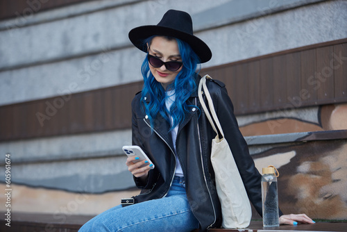 Stylish diverse person with dyed blue hair using a mobile app outdoor. Fashionable young woman sitting on a bench in the city center with a glass water bottle and tote bag on shoulder © hurricanehank