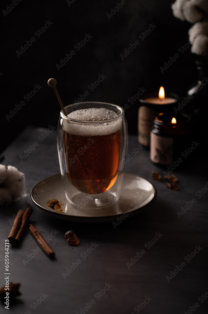 Freshly brewed black tea in a transparent glass with a steam on dark background with candles, cotton flowers and cinnamon sticks. Freshly brewed black tea in a transparent glass with a steam on dark background with candles, cotton flowers and cinnamon sticks.