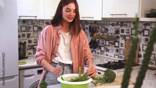 Young smiling beautiful woman in good mood selects vegetables for fresh vegan salad preparation for a healthy life in the kitchen of her home. Is taken vegetables and looks at them with real emotions photo