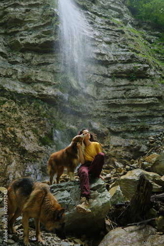 Young woman near waterfall with two dogs. Front view. Wanderlust, travel pets concept. Caucasian female traveler with australian and german shepherds enjoy nature while walking around the reserve.